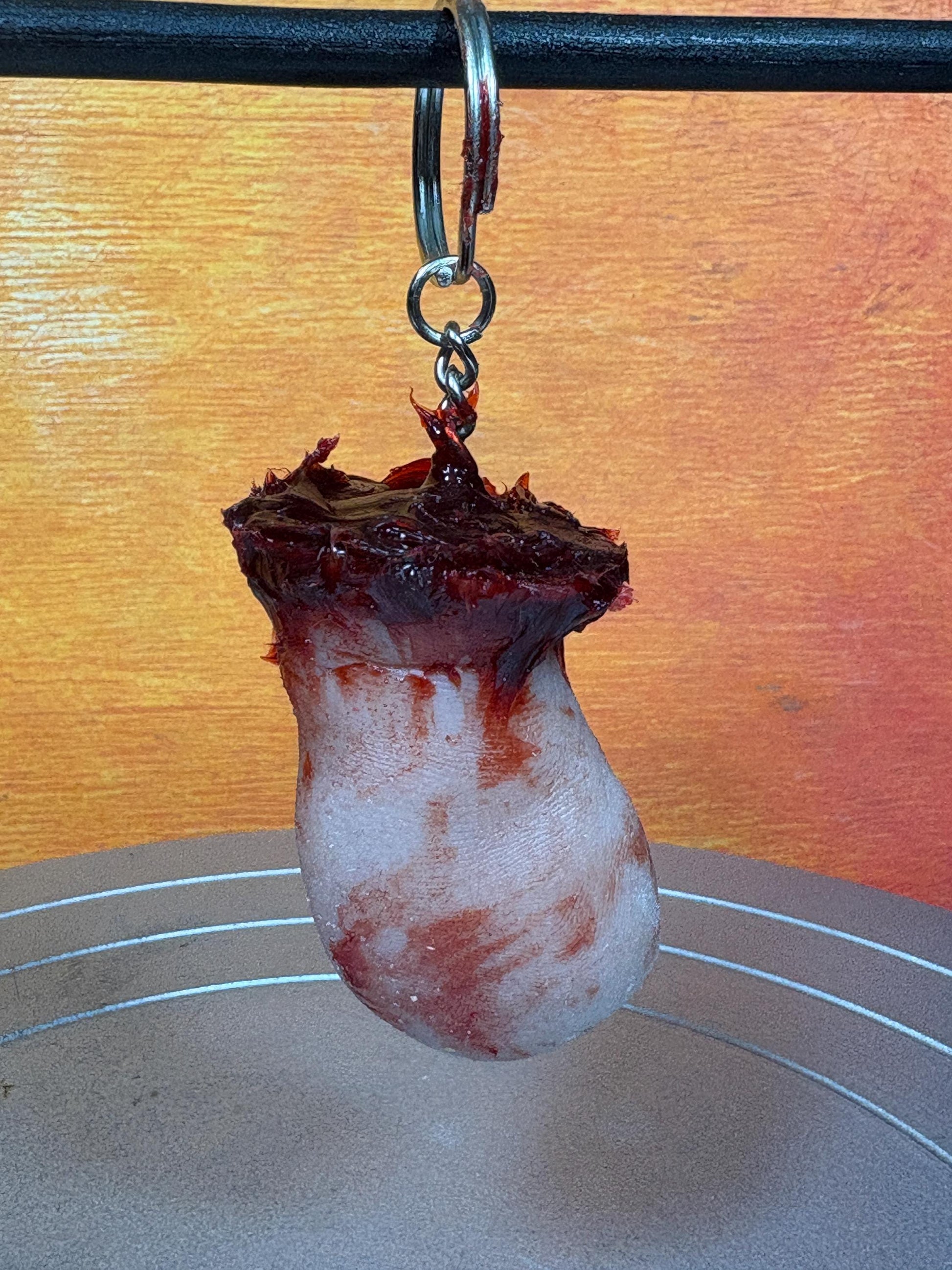 A white, blood-stained keychain charm resembling a severed finger or toe, topped with a dark, ragged, fleshy texture.